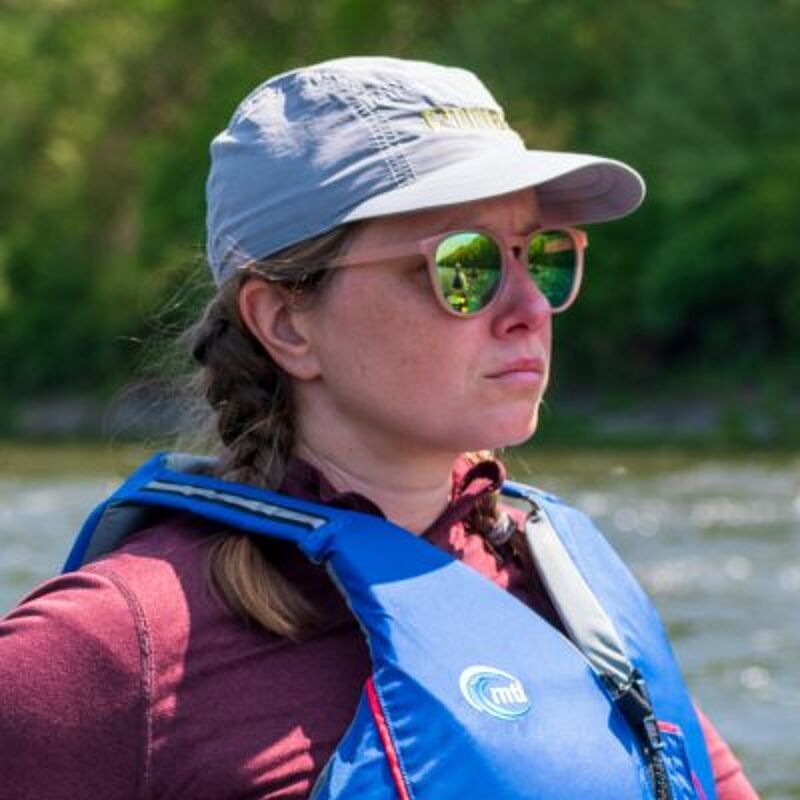 The image shows a woman wearing a cap, sunglasses, and a life jacket. She appears to be outdoors, possibly near a body of water, with trees visible in the background. The woman has a serious expression. The lighting suggests it is daytime.